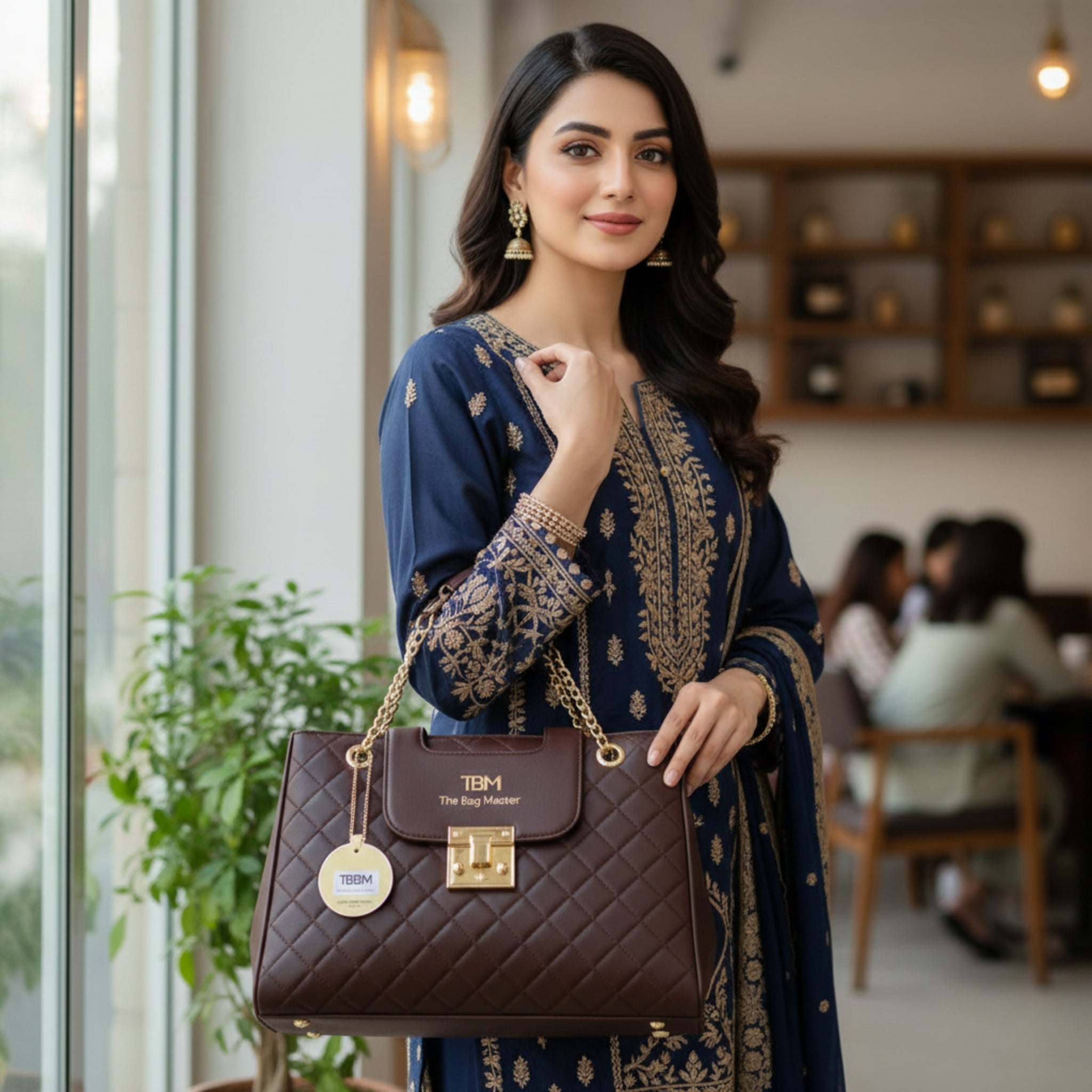 Woman holding a brown handbag with 'TBM' branding in an indoor setting.
